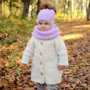 K&ouml;nnte beinhalten: Ein kleines Kind tr&auml;gt einen wei&szlig;en flauschigen Mantel, eine lila gestrickte M&uuml;tze mit einem braunen Bommel und einen lila gestrickten Schal. Das Kind steht in einem Wald mit heruntergefallenen Bl&auml;ttern auf dem Boden.