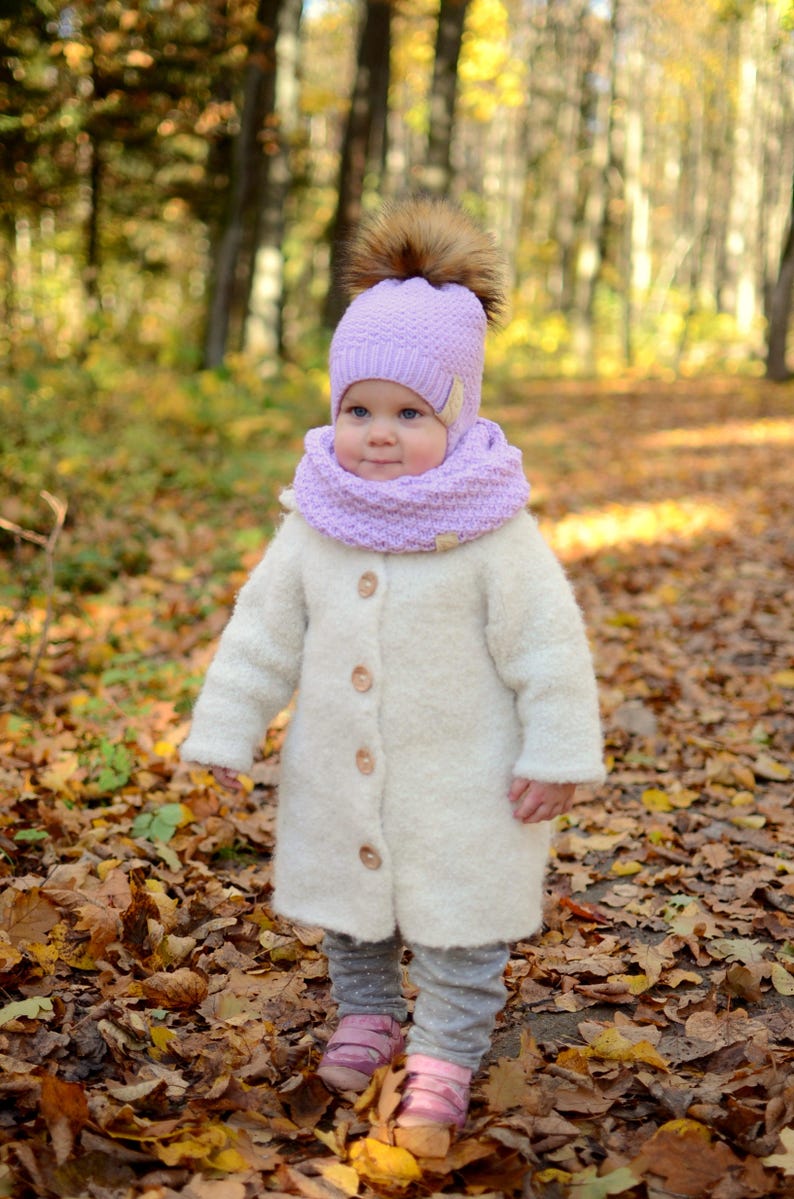 K&ouml;nnte beinhalten: Ein kleines Kind tr&auml;gt einen wei&szlig;en flauschigen Mantel, eine lila gestrickte M&uuml;tze mit einem braunen Bommel und einen lila gestrickten Schal. Das Kind steht in einem Wald mit heruntergefallenen Bl&auml;ttern auf dem Boden.