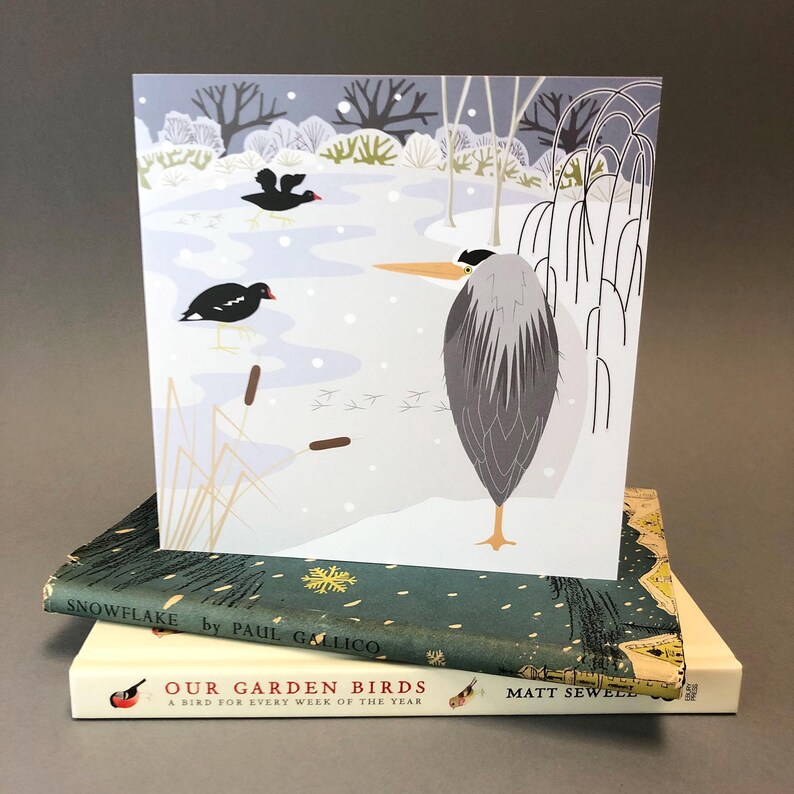 May include: A greeting card featuring a winter scene with a heron and two black birds in a snowy landscape. The card is propped up on two books, one titled "Snowflake" and the other "Our Garden Birds".