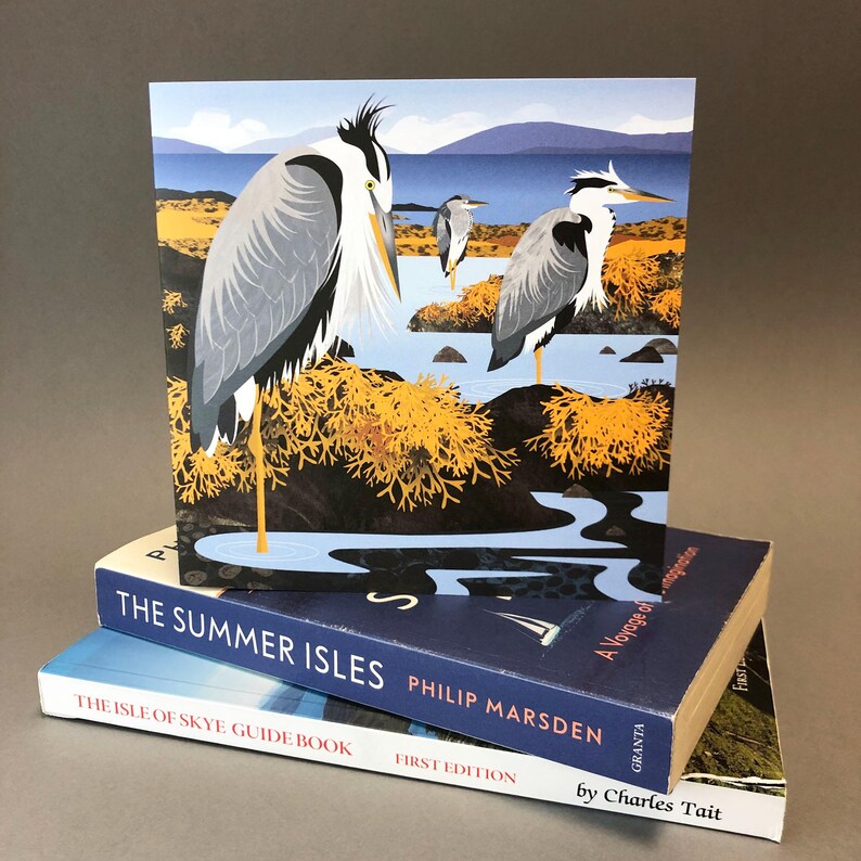 May include: A square art print featuring three herons in a coastal landscape with blue water, yellow foliage, and a blue sky. The print is propped up by two books: "The Summer Isles" and "The Isle of Skye Guide Book."