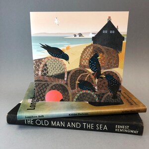 May include: A colorful illustration of a seaside scene with a black house, a beach, and a group of birds perched on top of stacked fishing baskets. The text "Longshore Drift" and "Katrina Porteous" is printed on the bottom of the card.