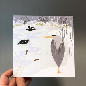 May include: A square art print featuring a winter scene with a gray heron and two black birds on a snowy landscape. The illustration includes bare trees, cattails, and bird tracks, with a soft, muted color palette.