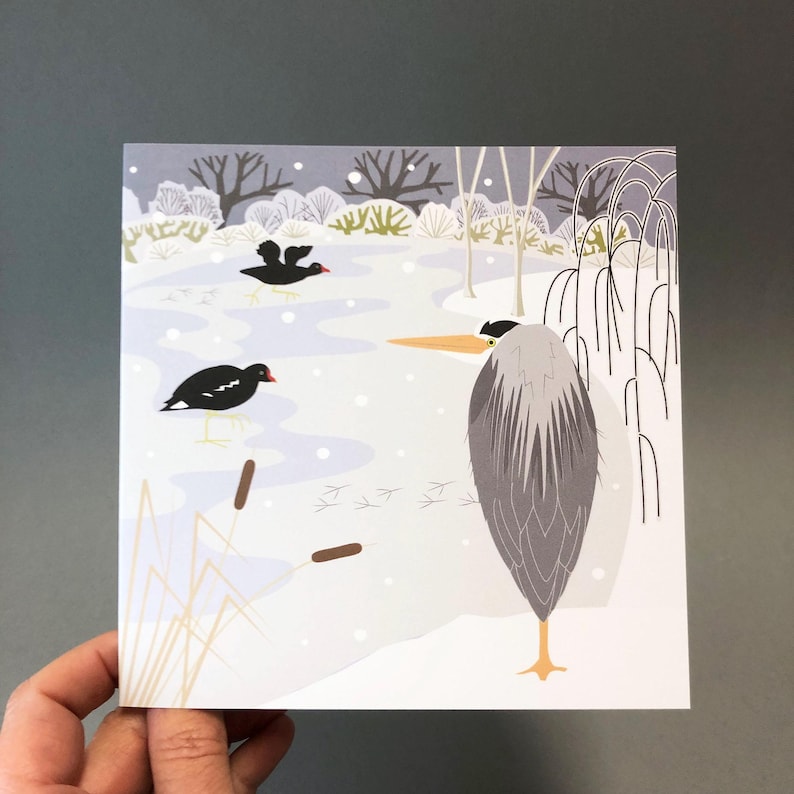 May include: A square art print featuring a winter scene with a gray heron and two black birds on a snowy landscape. The illustration includes bare trees, cattails, and bird tracks, with a soft, muted color palette.