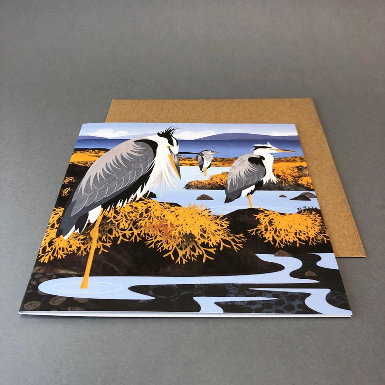 May include: Greeting card featuring a stylized illustration of three herons in a marshy landscape. The herons have gray and white feathers, long yellow legs, and stand near water and golden-yellow plants. The card is paired with a brown envelope.
