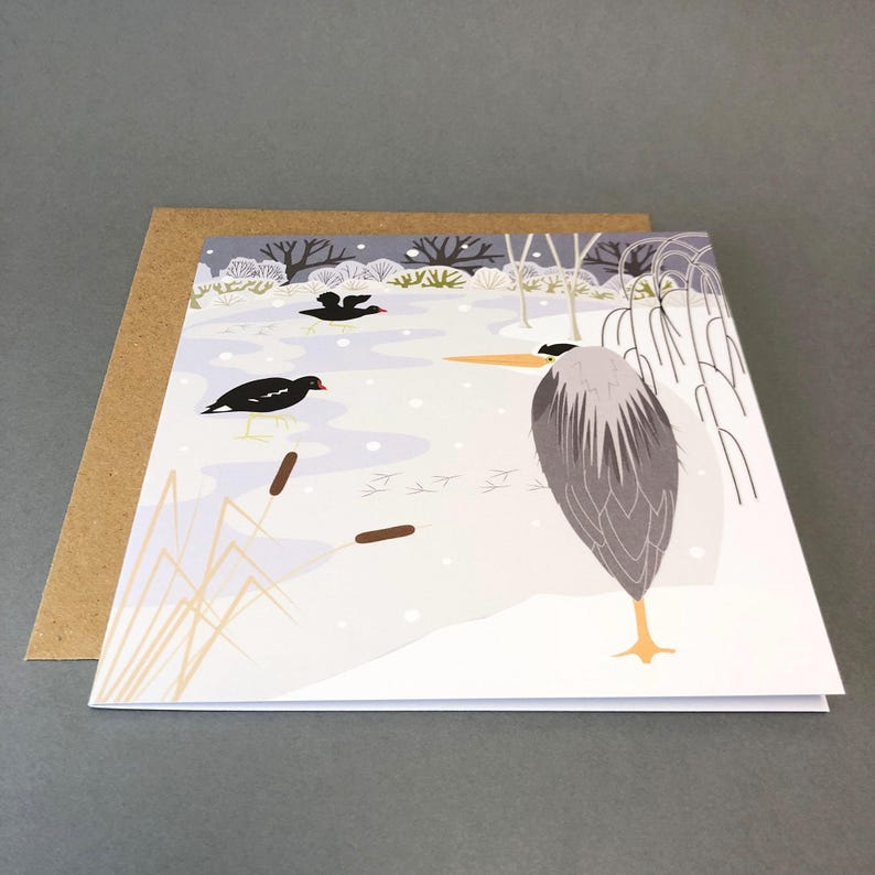 May include: A greeting card featuring a winter scene with a gray heron standing in the snow. The illustration includes black birds, cattails, and a snowy landscape. The card is paired with a brown envelope.