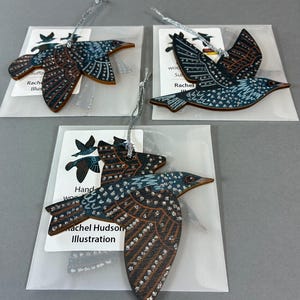 May include: Three wooden bird ornaments with silver hanging strings, each featuring dark blue and brown wings with white dotted patterns. The birds are packaged in clear plastic sleeves with white cards labeled Rachel Hudson Illustration.