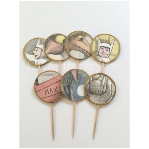 May include: A collection of eight round cupcake toppers featuring illustrations from the children's book "Where the Wild Things Are." Each topper has a gold glitter border and is attached to a wooden toothpick. One topper has the name "MAX" on it.