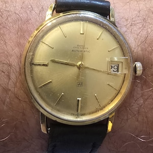 May include: A vintage gold-toned Seiko Fifth Avenue Automatic wristwatch with a black leather band. The watch face displays the date as the 29th. The watch has gold-colored hands and markers. The watch is on a wrist.