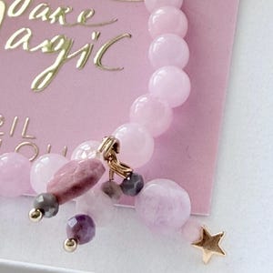 Könnte beinhalten: Ein rosa Perlenarmband mit einem goldenen Sternanhänger und verschiedenen Edelsteinakzenten. Das Armband hat eine Mischung aus runden und unregelmäßigen Perlen in Rosa- und Lilatönen. Im Hintergrund befindet sich eine rosa Karte mit dem goldenen Text "You are magic".