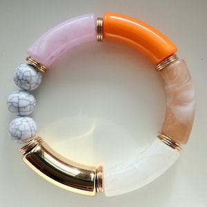 Könnte beinhalten: Ein farbenfrohes Armband mit einer Mischung aus Materialien und Farben. Das Armband besteht aus rosa, orangefarbenen, weißen und beigefarbenen Segmenten, die durch goldfarbene Metallakzente verbunden sind. Es hat auch weiß-grau marmorierte Perlen.