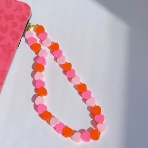Puede incluir: Una correa para teléfono de cuentas en forma de corazón rosa y naranja con detalles dorados.