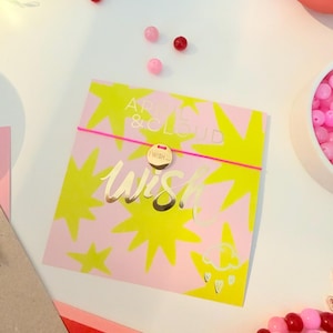 Könnte beinhalten: Eine rosa Karte mit gelben Sternenmustern und den goldenen Schriftzügen "APRIL & CLOUD" und "Wish". Ein goldener Anhänger mit "I Wish" ist an einem rosa Gummiband befestigt. Rosa und rote Perlen sind um die Karte herum verstreut.