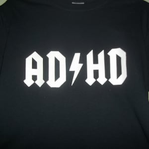 May include: Black t-shirt with the white text "AD⚡HD" in a bold, stylized font. The design features a lightning bolt replacing the "C" in a style reminiscent of a rock band logo. The shirt is a casual wear item.