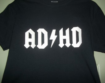 Peut inclure: T-shirt noir avec le texte blanc "AD⚡HD" dans une police de caractères audacieuse et stylisée. Le design présente un éclair remplaçant le "C" dans un style rappelant un logo de groupe de rock. Le t-shirt est un vêtement décontracté.