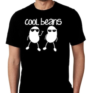 以下が含まれることがあります： 黒いTシャツ。白い文字で「cool beans」と書かれ、サングラスをかけた2つの白い豆のグラフィックが描かれています。豆は棒人間の腕と脚を持ち、1つは手を振っています。