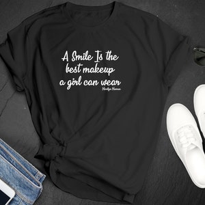 Pode incluir: T-shirt preto com a citação "A Smile Is the best makeup a girl can wear" em escrita branca, atribuída a Marilyn Monroe. A camisa é estilizada com óculos de sol, tênis brancos e jeans com um smartphone e fones de ouvido.