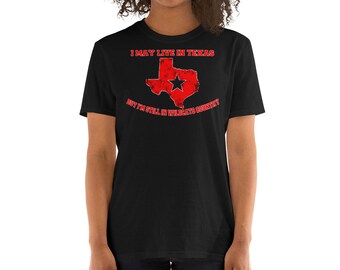 Op de afbeelding: Zwart T-shirt met een verweerde rode Texas-omtrek en een zwarte ster. De tekst luidt "I MAY LIVE IN TEXAS" boven de afbeelding en "BUT I'M STILL IN WILDCATS COUNTRY" eronder. Een casual kledingstuk.