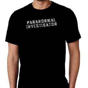 May include: Black t-shirt featuring the text "PARANORMAL INVESTIGATOR" in a distressed white font. The t-shirt has a crew neck and is made of a soft material. The image shows the upper body of a person wearing the shirt.