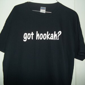 May include: Black t-shirt with the white text "got hookah?" printed across the front. The shirt is hanging on a hanger against a white wall. The t-shirt is a short-sleeved, crew neck style.