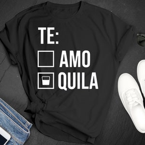 May include: Black t-shirt with white text that reads "TE: ☐ AMO ☐ QUILA". The shirt is styled with blue jeans, white trainers, and sunglasses. A smartphone with white earbuds is also visible.