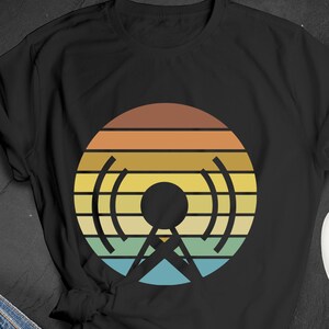 Peut inclure: T-shirt noir avec un motif rétro de tour radio. Le design présente une forme circulaire avec des rayures horizontales marron, orange, jaune, vert et bleu. L'antenne est noire.