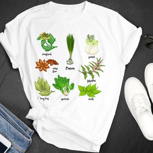 Peut inclure: T-shirt blanc avec des illustrations colorées d'herbes et d'épices, dont la moutarde, l'anis étoilé, la feuille de laurier, les épinards, l'oignon, le fenouil, le sésame et la menthe. Les noms de chaque herbe sont imprimés en dessous. Lunettes de soleil et chaussures blanches également visibles.