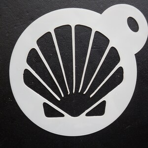 May include: A white plastic stencil with a seashell design. The stencil is circular with a handle and cut-out lines forming the seashell pattern. The background is black.