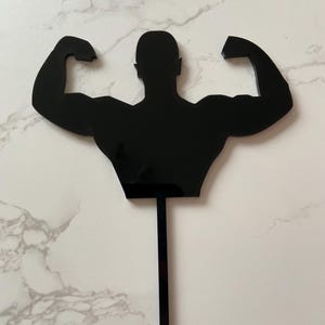 Könnte beinhalten: Schwarzer Acryl-Kuchenaufsatz mit einer Silhouette eines Bodybuilders, der seine Muskeln anspannt. Die Figur hat die Arme erhoben und zeigt muskulöse Bizeps. Der Aufsatz hat einen langen, dünnen Stiel zum Einstecken in einen Kuchen. Ideal für Fitness-Themenfeiern.