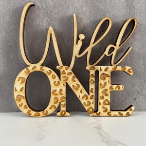 May include: Wooden sign reading "Wild One". The word "ONE" features a leopard print design. The sign is made of light-coloured wood and is likely used as a party decoration for a first birthday.