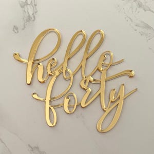 May include: A gold-coloured acrylic cake topper featuring the words hello forty in a cursive script font. The surface of the lettering is reflective and smooth, creating a metallic appearance with clean, sharp edges throughout the text design.