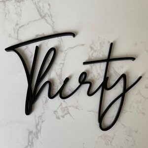 May include: A black acrylic sign featuring the word Thirty written in a continuous cursive script font. The lettering has a smooth, glossy finish and is designed as a single connected piece of decor with a distinct, flowing calligraphic style.