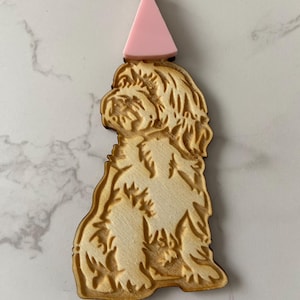 May include: A light brown wooden dog-shaped decoration, wearing a pink party hat. The dog has detailed fur texture. The pink party hat is attached to the top of the dog's head, adding a festive touch.