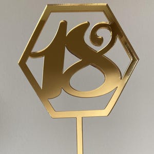 以下が含まれることがあります： 六角形の金色の鏡面ケーキトッパー。装飾的なフォントで18の数字が書かれています。