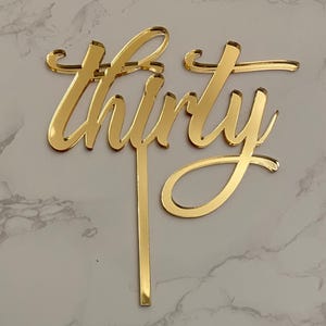 May include: A gold-coloured cake topper spelling out the word "thirty" in a cursive font. The topper is made of a reflective material and is designed to be inserted into a cake. The background is a light grey and white marble pattern.