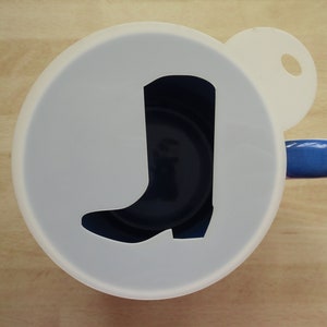 May include: A white stencil featuring a cowboy boot cutout, positioned over a dark blue mug. The mug has a blue handle. The design is centrally placed within the stencil, creating a visual contrast.
