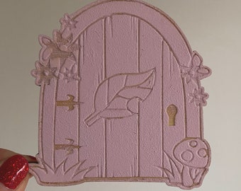 May include: A pink wooden fairy door with a leaf design, a keyhole, and a mushroom at the bottom. The door is decorated with carved flowers and vines.