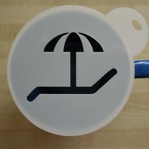 Peut inclure: Une tasse en céramique blanche avec une poignée bleue et un pochoir noir représentant une chaise longue et un parasol de plage.