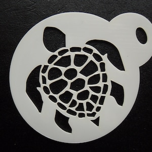 May include: A white plastic stencil with a sea turtle design. The turtle is in the centre of a circular stencil with a handle. The turtle's shell has a detailed, geometric pattern. The stencil is likely used for crafts or decorating.