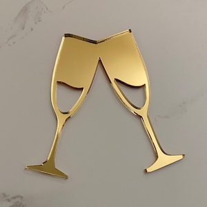 May include: Two gold-coloured champagne flutes, raised in a toast, are shown against a white marble background.