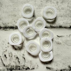 May include: A collection of white, square-shaped bottle stoppers with a circular, recessed center. The stoppers are made of a translucent material and are arranged on a textured, light-colored surface. The stoppers are designed to seal bottles.