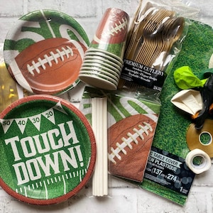 May include: A football-themed party supplies set featuring plates, cups, napkins, cutlery, balloons, and a green table cover. The plates have a football and field design, with one plate reading "Touch Down!"