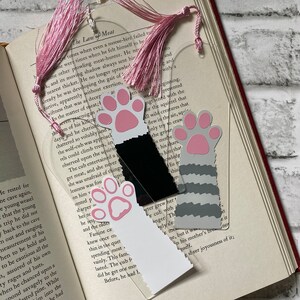 May include: Three clear acrylic bookmarks with paw print designs. The bookmarks feature pink and white paw prints, a black and white striped paw print, and a black paw print. The bookmarks are decorated with pink and white tassels.