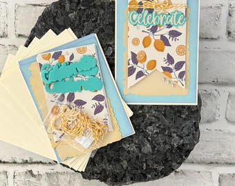 May include: A card-making kit with a lemon theme. The kit includes a card with the word "Celebrate", a banner with lemon illustrations, and other crafting supplies. The color scheme is light blue, yellow, and purple.