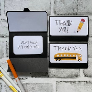 May include: Black gift card holder with a white insert that reads "INSERT YOUR GIFT CARD HERE." Two "Thank You" cards are also shown, one with a pencil graphic and the other with a school bus graphic. Crayola markers are in the foreground.