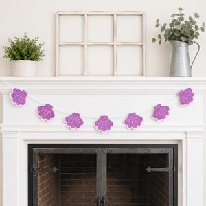 May include: A decorative garland featuring glittery purple seashell shapes with a light pink border, strung on a white string. The garland is displayed above a fireplace, suggesting a coastal or mermaid-themed decoration.