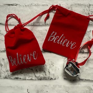 May include: Two red velvet drawstring bags with the word "Believe" in silver glitter script. One bag has a small star next to the word. A silver bell with a red ribbon is attached to one bag. The bags are on a white brick background.