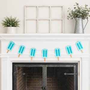 May include: A garland of blue popsicle decorations with two vertical stripes on each, strung on a white string. The garland is hanging above a fireplace. The background includes a white window frame, a plant in a pot, and a metal jug with greenery.