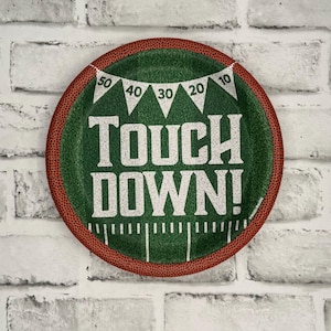 May include: A round paper plate with a football field design. The center is green with white yard lines and the words "TOUCH DOWN!" in large white letters. A banner with numbers 10, 20, 30, 40, and 50 is at the top. The border is brown.