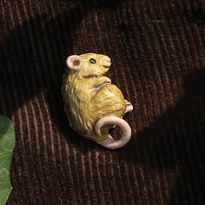 Puede incluir: Pequeña figura detallada de un ratón en posición sentada. El ratón es de color amarillo claro con cola y orejas rosas. Descansa sobre una tela marrón, similar a la pana, con hojas verdes de hiedra en el fondo.
