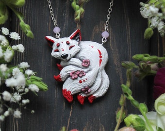 Op de afbeelding: Een witte keramische vos-hanger met rode en roze accenten, gedragen aan een zilveren kettingketting. De hanger is omringd door witte en roze bloemen.